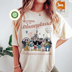 May include: Cream-colored t-shirt featuring a Disneyland graphic. The design includes the text "CALIFORNIA Disneyland EST. 1955" above an illustration of the castle, characters, and the phrase "The happiest place on earth."