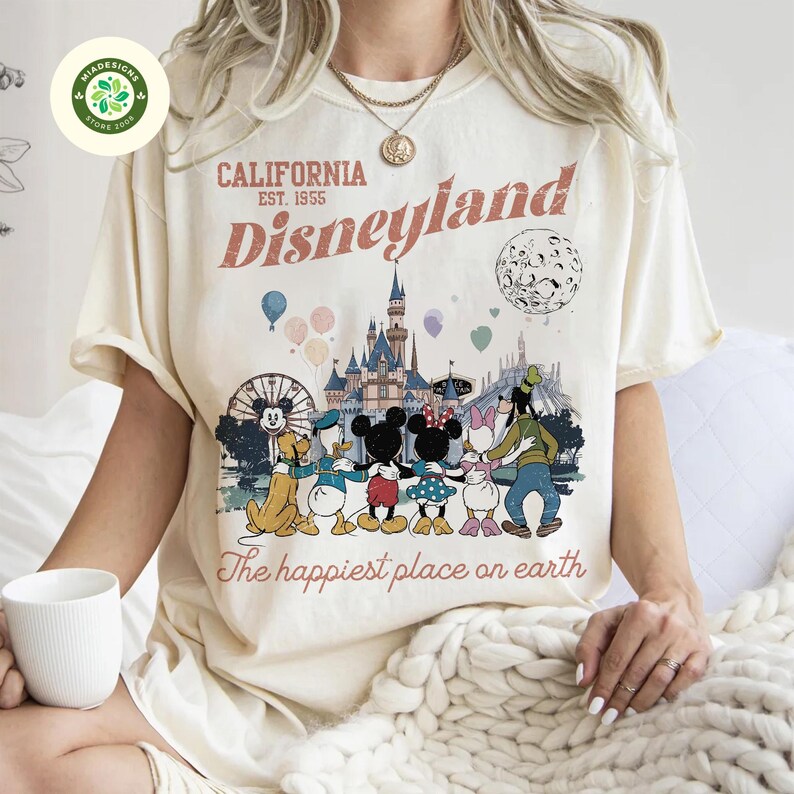 May include: Cream-colored t-shirt featuring a Disneyland graphic. The design includes the text "California Disneyland Est. 1955" and a colorful illustration of Disney characters and the castle. The phrase "The happiest place on earth" is also printed on the shirt.