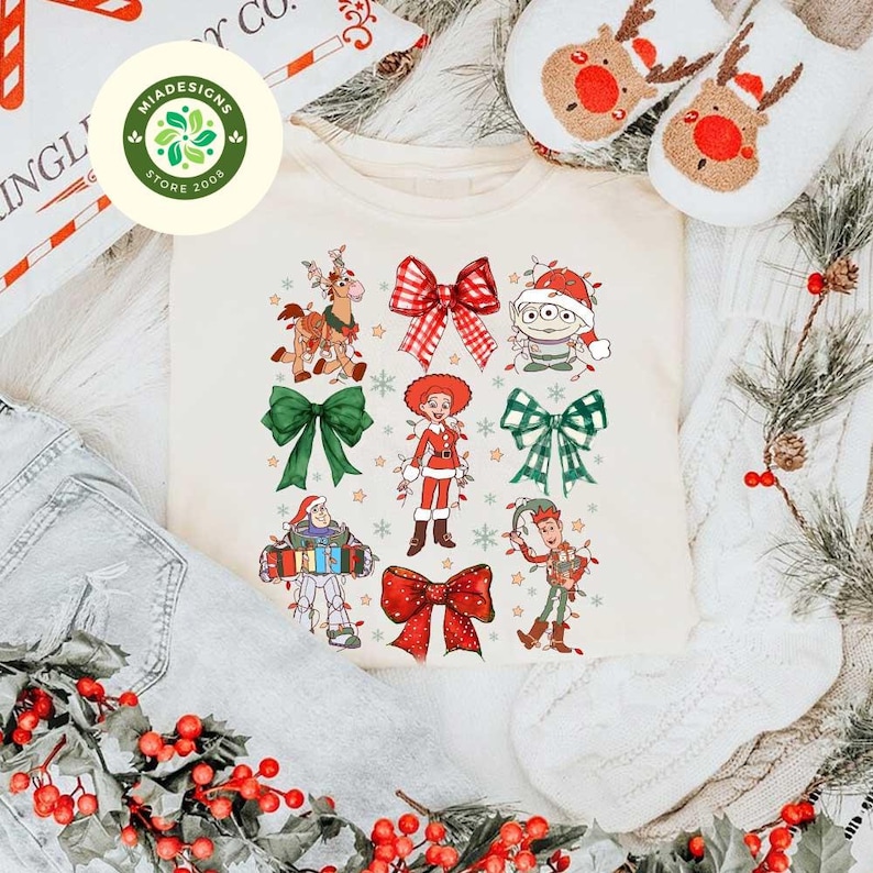 May include: Cream-colored crew neck t-shirt featuring a festive design with cartoon characters and bows. The design includes characters like Woody, Buzz Lightyear, and an alien, all adorned with Christmas lights and bows. The shirt is surrounded by holiday decor.