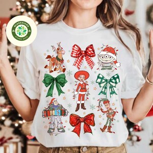 May include: White t-shirt featuring Christmas-themed cartoon characters and bows. The design includes characters like Buzz Lightyear and Little Green Men, with festive bows in red and green. The shirt has a round neck.