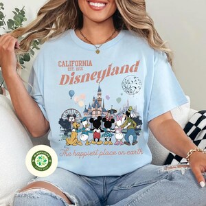 May include: Light blue t-shirt with a Disneyland graphic. The graphic features Mickey Mouse, Minnie Mouse, Donald Duck, Daisy Duck, and Goofy in front of a castle, Ferris wheel, and balloons. Text reads "California Disneyland Est. 1955" and "The happiest place on earth."