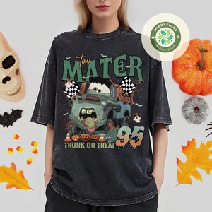 May include: A dark gray t-shirt featuring a Halloween-themed graphic of Tow Mater from the movie Cars. The design includes pumpkins, ghosts, and the text "Tow Mater Trunk or Treat 95".