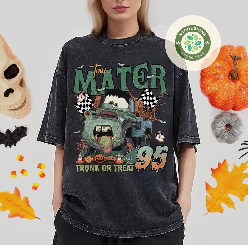 May include: A dark gray t-shirt featuring a Halloween-themed graphic of Tow Mater from the movie Cars. The design includes pumpkins, ghosts, and the text "Tow Mater Trunk or Treat 95".
