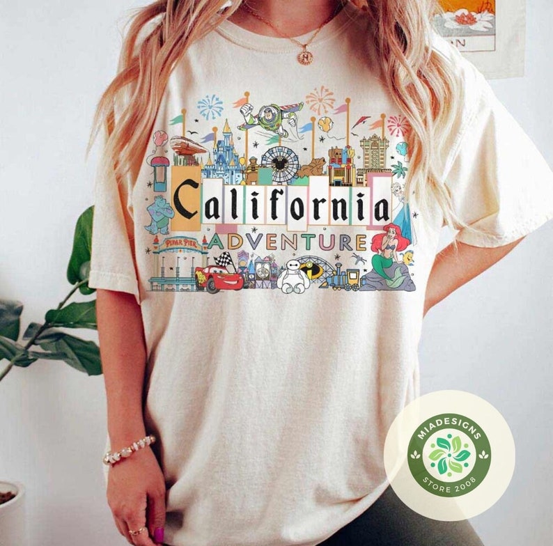 May include: A beige t-shirt with a colorful graphic of the California Adventure theme park. The graphic includes iconic attractions, characters, and the words "California Adventure".