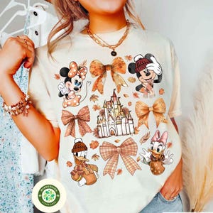 May include: Cream-colored t-shirt featuring cartoon characters Mickey Mouse, Minnie Mouse, Donald Duck, and Daisy Duck, along with autumn-themed bows, leaves, and a castle. The shirt has a round neck and short sleeves.