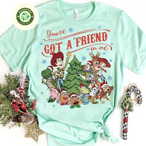 May include: A light green t-shirt featuring a festive graphic of characters from the Toy Story movie. The design includes Woody, Buzz Lightyear, and other characters around a Christmas tree, with the text "You've Got A Friend in Me".