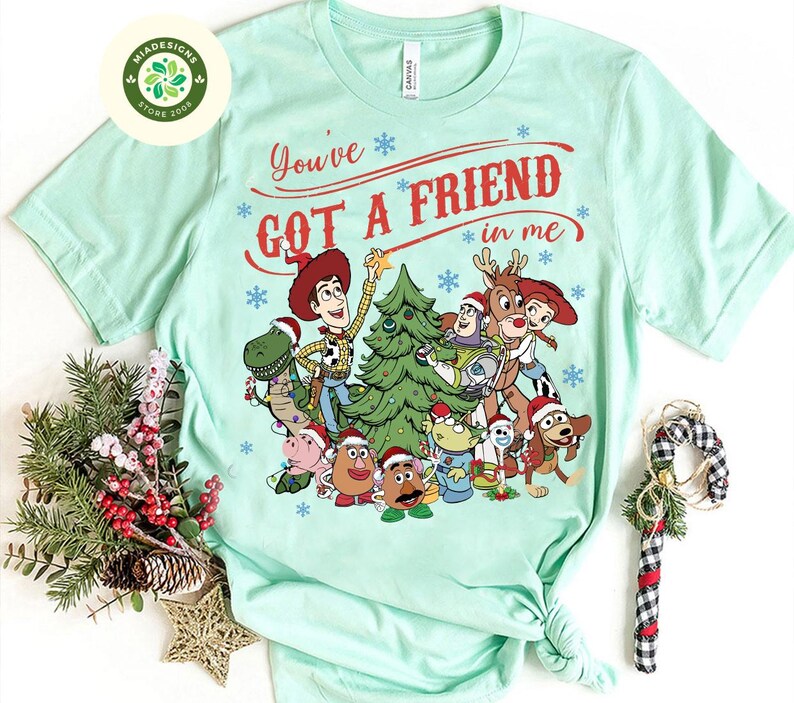 May include: A light green t-shirt featuring a festive graphic of characters from the Toy Story movie. The design includes Woody, Buzz Lightyear, and other characters around a Christmas tree, with the text "You've Got A Friend in Me".