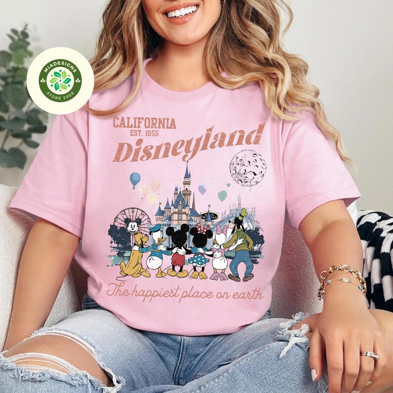May include: Pink t-shirt with a Disneyland graphic. The design features the Disneyland logo, the text "California Est. 1955 Disneyland", and a cartoon illustration of Disney characters. The phrase "The happiest place on earth" is also included.