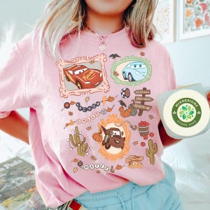 以下が含まれることがあります： ピンク色のTシャツ。映画「カーズ」のキャラクター、ライトニング・マックィーンやメーターなどの漫画イラストが、装飾的なフレームの中に描かれています。カボチャ、サボテン、標識などのグラフィックも含まれています。緑色のロゴが入ったステッカーが手に持たれています。