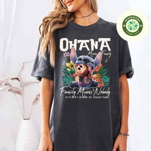 May include: Dark gray t-shirt featuring a graphic of the character Stitch from Lilo & Stitch. The design includes the words "Ohana Means Family" and "Family Means Nobody Gets Left Behind or Forgotten". The image also includes colorful flowers.