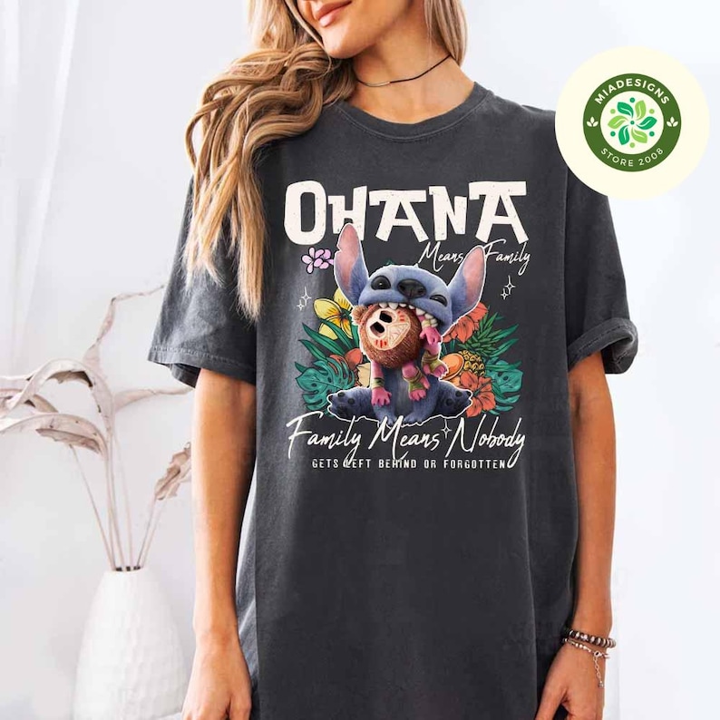 May include: Dark gray t-shirt featuring a graphic of the character Stitch from Lilo & Stitch. The design includes the words "Ohana Means Family" and "Family Means Nobody Gets Left Behind or Forgotten". The image also includes colorful flowers.