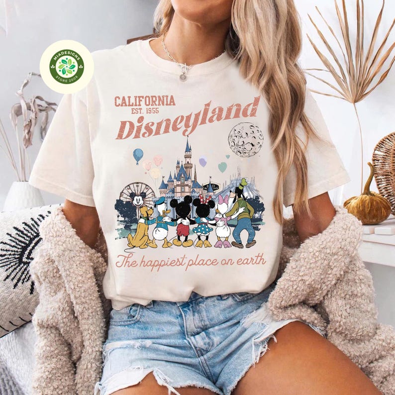 May include: Cream-colored t-shirt featuring a Disneyland graphic. The design includes the text "CALIFORNIA EST. 1955 Disneyland" above a colorful illustration of Disney characters and the castle. The phrase "The happiest place on earth" is printed below.