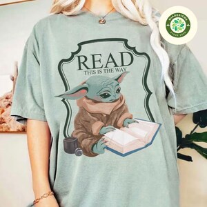 May include: A light green t-shirt with a graphic of a green alien character reading a book. The graphic is surrounded by a green frame with the text "READ THIS IS THE WAY".