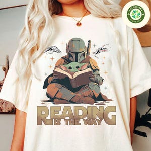 May include: A white t-shirt with a graphic of a Mandalorian character holding a book with a small green alien character sitting on the book. The text "Reading is the way" is printed below the graphic.