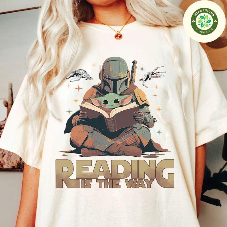 May include: A white t-shirt with a graphic of a Mandalorian character holding a book with a small green alien character sitting on the book. The text "Reading is the way" is printed below the graphic.