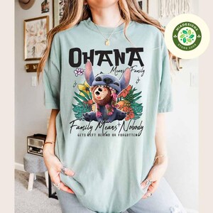 May include: A light blue t-shirt with the words "OHANA Means Family" in black. The shirt features a graphic of Stitch from Lilo & Stitch, surrounded by tropical flowers and the text "Family Means Nobody Gets Left Behind or Forgotten."