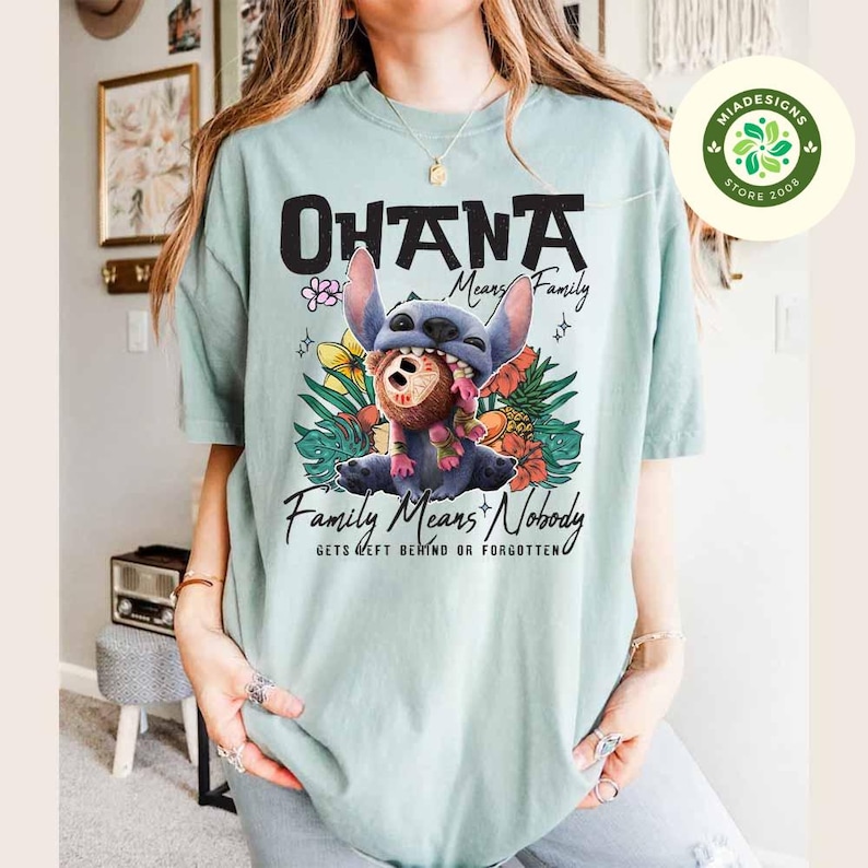 May include: A light blue t-shirt with the words "OHANA Means Family" in black. The shirt features a graphic of Stitch from Lilo & Stitch, surrounded by tropical flowers and the text "Family Means Nobody Gets Left Behind or Forgotten."
