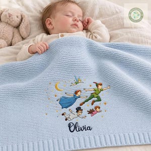 May include: A light blue knitted baby blanket featuring an embroidered design of Peter Pan characters flying with a yellow crescent moon and stars. The name Olivia is embroidered in dark blue script below the characters. A plush toy is visible.
