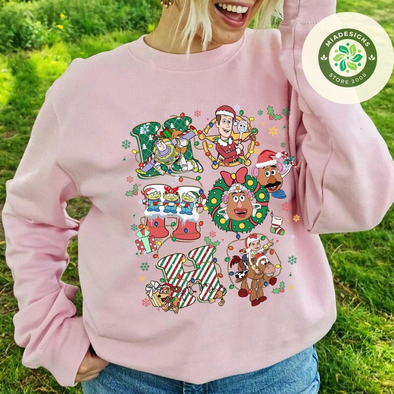 Puede incluir: Sudadera rosa claro con un dise&ntilde;o navide&ntilde;o colorido. El dise&ntilde;o incluye varios personajes de dibujos animados, luces navide&ntilde;as y la frase "Ho Ho Ho" en una fuente festiva. La sudadera tiene mangas largas y cuello redondo.