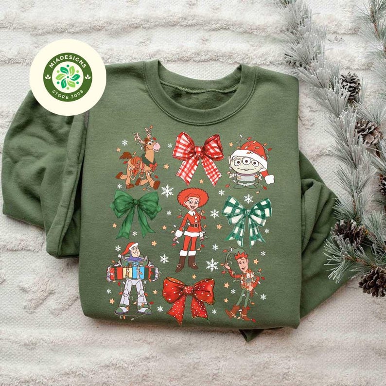 May include: An olive green sweatshirt featuring a festive design with cartoon characters and Christmas bows. The graphic includes characters like Woody, Buzz Lightyear, and the Little Green Men, all adorned with holiday elements. The design is surrounded by snowflakes.