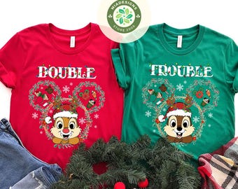 May include: Two Christmas-themed t-shirts, one red and one green. The red shirt says "DOUBLE" and the green shirt says "TROUBLE". Each shirt features a Chip and Dale graphic wearing a Santa hat, inside a Mickey Mouse head-shaped wreath.
