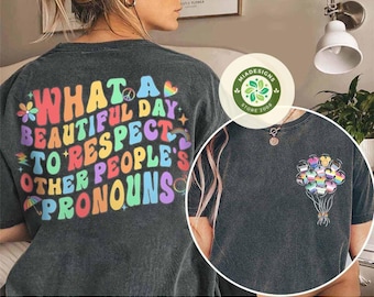 May include: Two black t-shirts with rainbow text and graphics. The first shirt says "What a beautiful day to respect other people's pronouns". The second shirt has a graphic of rainbow balloons.