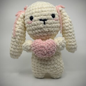 May include: A handmade, cream-colored crochet bunny with pink accents. The bunny has black eyes, pink cheeks, and a pink heart. The bunny's ears have pink trim, and it is holding a pink heart.