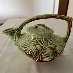 Pode incluir: Uma chaleira de cerâmica verde e marrom com um design de folhas. A chaleira tem uma alça e um bico.