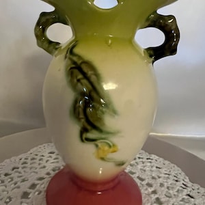Può includere: Un vaso in ceramica vintage con un design floreale verde e bianco. Il vaso ha due manici e una base rosa.