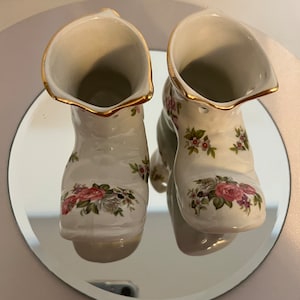 Może przedstawiać: Para białych porcelanowych wazonów w kształcie butów z kwiatowymi wzorami. Wazy mają złotą obwódkę wokół górnej części i są ozdobione różowymi i białymi kwiatami.