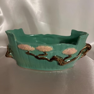 May include: A teal ceramic planter with a brown tree branch design and pink flowers. The planter has a unique shape with a wide opening and a narrow base.