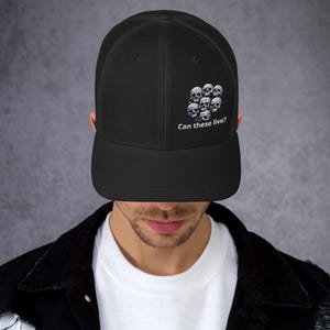 May include: A black mesh trucker hat with a white embroidered design of skulls and the text "Can these live?"