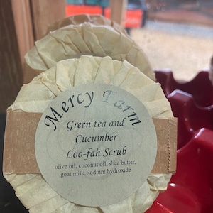 May include: A round, white, paper-wrapped soap bar with the text "Mercy Farm" in a circle. The soap is labeled "Green tea and Cucumber Loo-fah Scrub" and lists the ingredients: olive oil, coconut oil, shea butter, goat milk, and sodium hydroxide.
