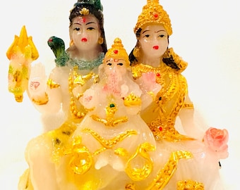 May include: White marble statue of a Hindu deity family, Shiva, Parvati, and Ganesha. The figures are seated together, with Shiva holding a trident and Parvati holding a lotus flower. The statue is decorated with gold accents and is set on a black base.