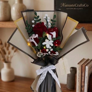 Puede incluir: Un ramo artesanal con rosas de tela rojas y blancas, flores blancas y detalles verdes, envuelto en papel negro y beige con una cinta de encaje blanco. De NamasteCraftsCo.