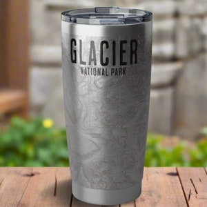 May include: A stainless steel tumbler with a map design of Glacier National Park. The words "GLACIER NATIONAL PARK" are printed in black. The tumbler has a black lid and is sitting on a wooden surface.