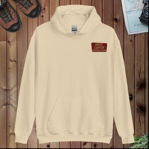 May include: A cream-colored hoodie with a front pocket and a hood. The upper left chest features a small rectangular patch with the text "BRYCE CANYON NATIONAL PARK" in red and white. The hoodie is made of a soft material.