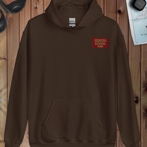 May include: A brown hoodie with a front pocket and a small rectangular patch on the chest. The patch reads "SEQUOIA NATIONAL PARK" in white on a red background. The sweatshirt is made of a soft material. The hoodie has a drawstring.