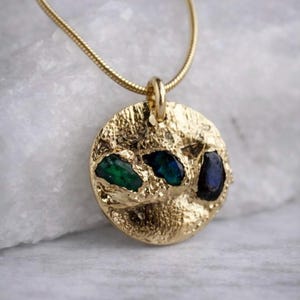 May include: A gold-colored pendant necklace with a textured, circular pendant. The pendant features three inset gemstones in shades of green and blue. The necklace chain is a thin, gold-colored snake chain.