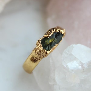 Könnte beinhalten: Ein goldfarbener Ring mit einem rauen, strukturierten Band und einem dunkelgrünen und gelben Edelstein. Der Edelstein ist rechteckig und in einer strukturierten Goldfassung gefasst. Der Ring wird auf einer weißen Oberfläche präsentiert.