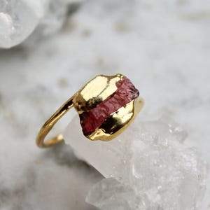 Op de afbeelding: Een gouden ring met een ruwe roze edelsteen die in de band is gezet. De ring is van goud en heeft een eenvoudig ontwerp.