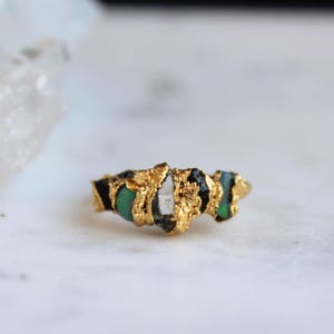 Op de afbeelding: Een goudkleurige ring met een getextureerde band, met ruwe edelstenen in groen, zwart en wit. Het ontwerp van de ring is natuurlijk en organisch, waarbij de unieke vormen en kleuren van de stenen worden benadrukt.