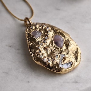 May include: A gold-toned pendant necklace featuring an irregular, textured, organic-shaped metal surface embedded with several small, light purple and iridescent stone fragments, suspended from a thin, matching gold-toned chain.