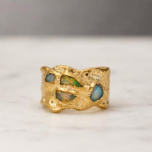 Puede incluir: Un anillo ancho de oro con una superficie orgánica y texturizada que presenta cuatro ópalos irregulares incrustados. El metal tiene un acabado rugoso y martillado, y los ópalos muestran destellos de colores verde, naranja y azul en su interior.