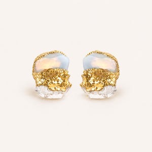 May include: A pair of stud earrings featuring a gold-colored, textured setting. Each earring has a light blue, oval-shaped stone at the top and a clear, faceted stone at the bottom. The earrings are displayed on a white background.