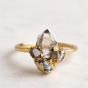 May include: A gold ring featuring a large, clear crystal with a pointed top and several smaller, gray crystals clustered around it. The ring has a textured gold band and a gold-colored setting.