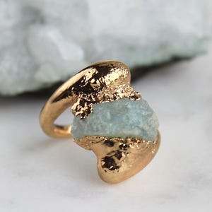 May include: A gold-colored ring featuring a rough, light blue gemstone. The ring has a textured, organic design, with the gemstone partially encased in the gold setting. The ring is set against a blurred background.