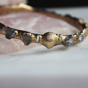 May include: A gold-colored bangle bracelet featuring a seashell and rough-cut gemstones. The bracelet has a textured, organic design with a seashell in the center and several gemstones. The background is a soft pink and white.