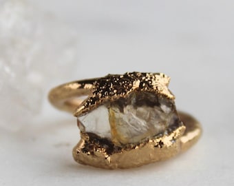 May include: A gold-colored ring featuring a rough-cut, clear gemstone. The gemstone is encased in a textured gold setting. The ring's band is smooth and polished, contrasting with the textured gemstone setting. The ring is set against a soft, neutral background.