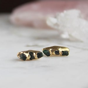 Puede incluir: Un par de pendientes de oro, cada uno con tres pequeñas piedras de esmeralda en bruto de color verde oscuro engastadas horizontalmente en una barra de oro pulido. El diseño presenta un acabado metálico texturizado y orgánico.